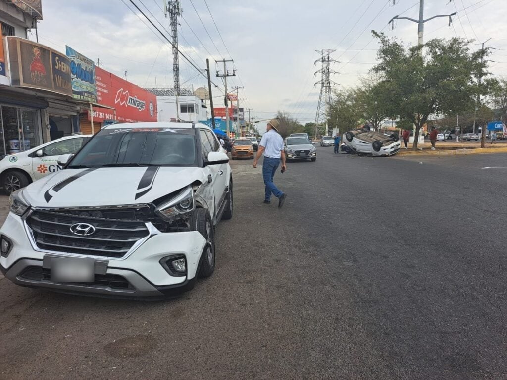 Una camioneta blanca chocada, algunos carros al fondo y un carro color blanco volcado con los neumáticos hacia arriba a un lado de un palo de un árbol