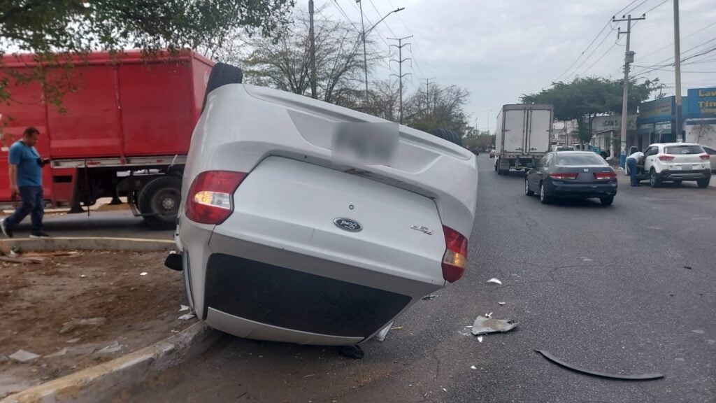 Carro color blanco volcado con los neumáticos hacia arriba, la cola de un camión rojo y algunos carros de fondo