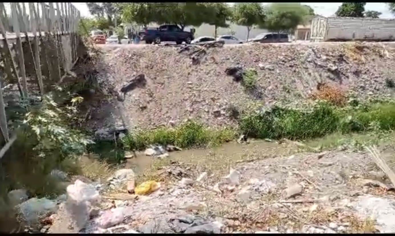 Canal con poca agua y mucha basura, al fondo carros y barandal de fierro de un puente que cruza la vía hidráulica