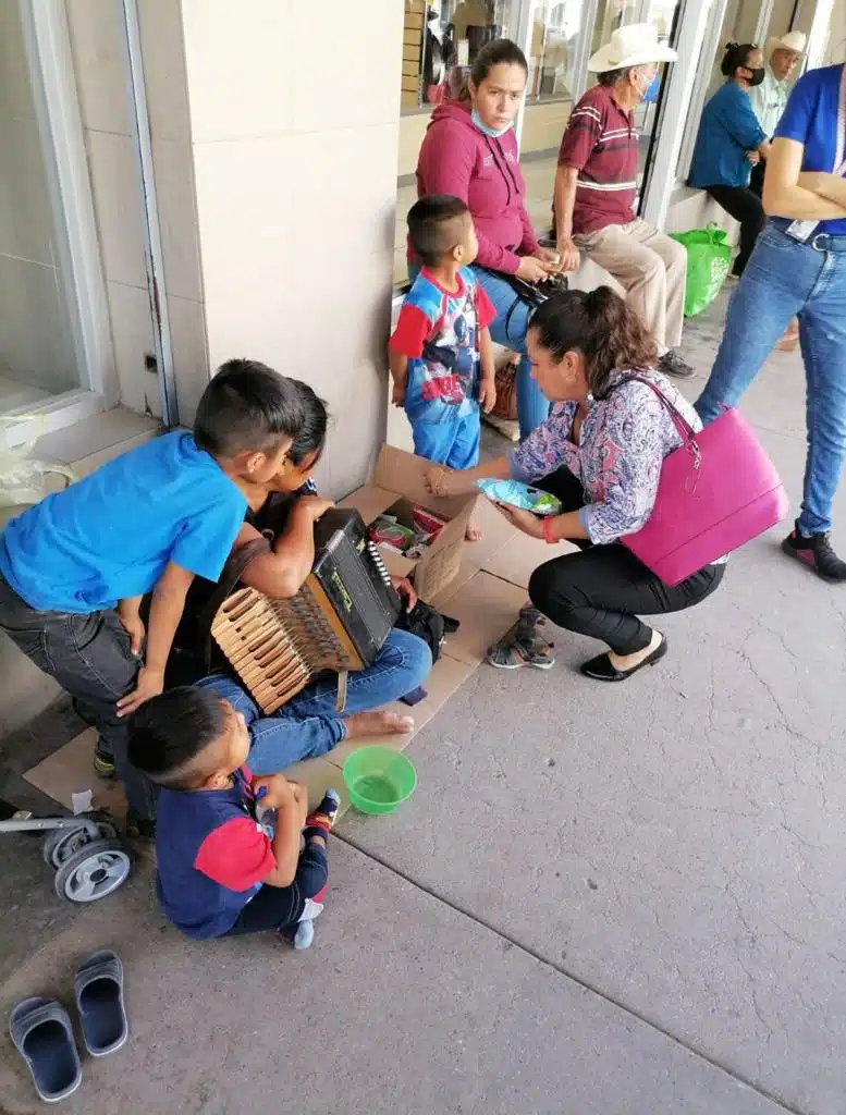 Una señora con un acordeón y un niño sentados en el piso, 2 niños parados y una señora en cuclillas viendo lo que hay en un cartón