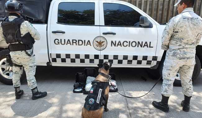 2 personas y un perro mirando hacía unos paquetes y una camioneta de la Guardia Nacional