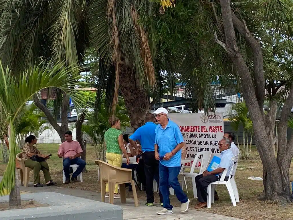 Varias personas sentadas, tres paradas y una lona atrás