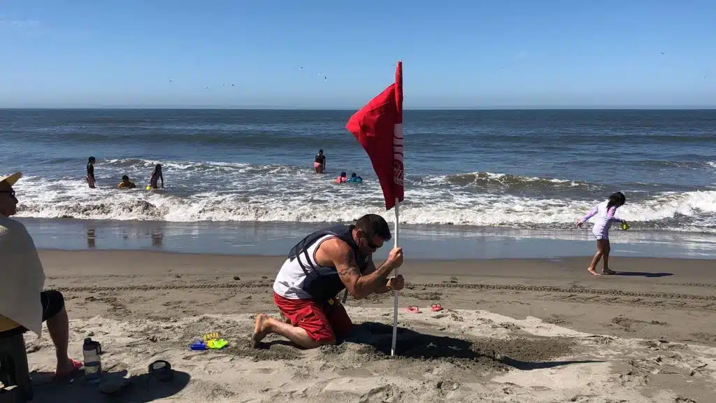 Una persona poniendo una bandera roja en la arena, otra persona viéndola y varias personas bañándose en el mar