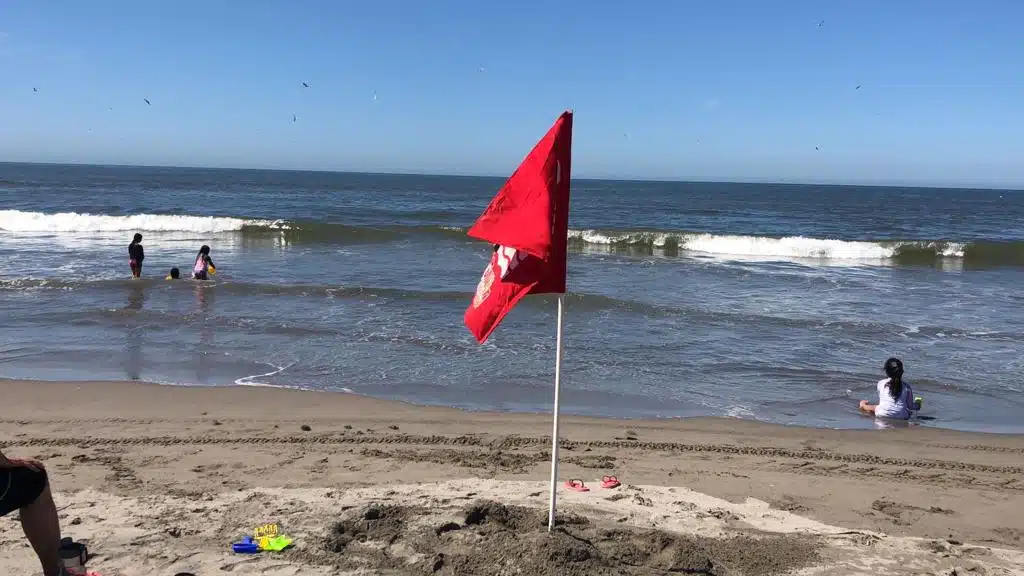 Una bandera roja en la arena y varias personas bañándose en el mar