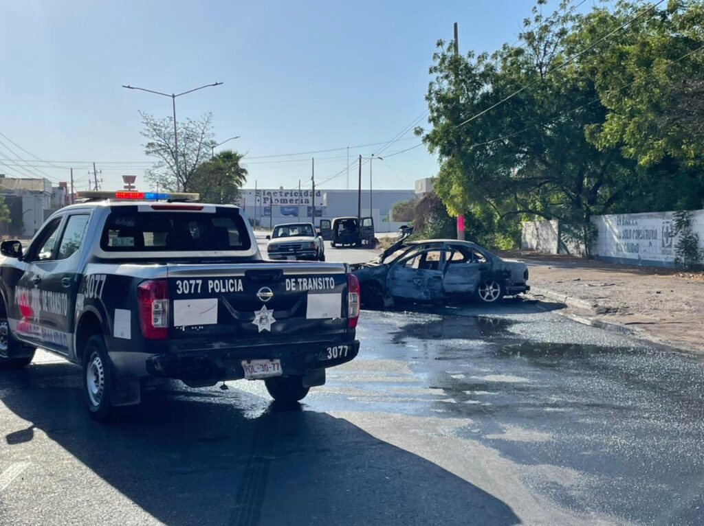 Patrulla de la Policía Municipal en primer plano, de fondo el vehículo Nissan Sentra calcinado tras incendiarse luego de un choque en calles de Culiacán