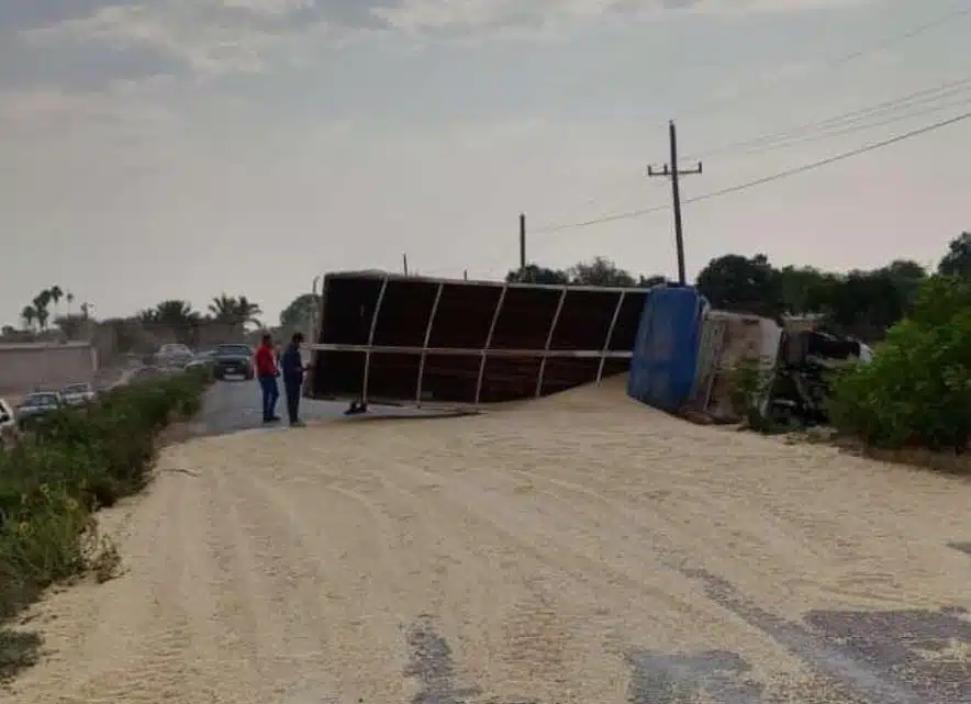 2 personas paradas en frente de un torton volcado en una carretar con granos de maiz en el suelo