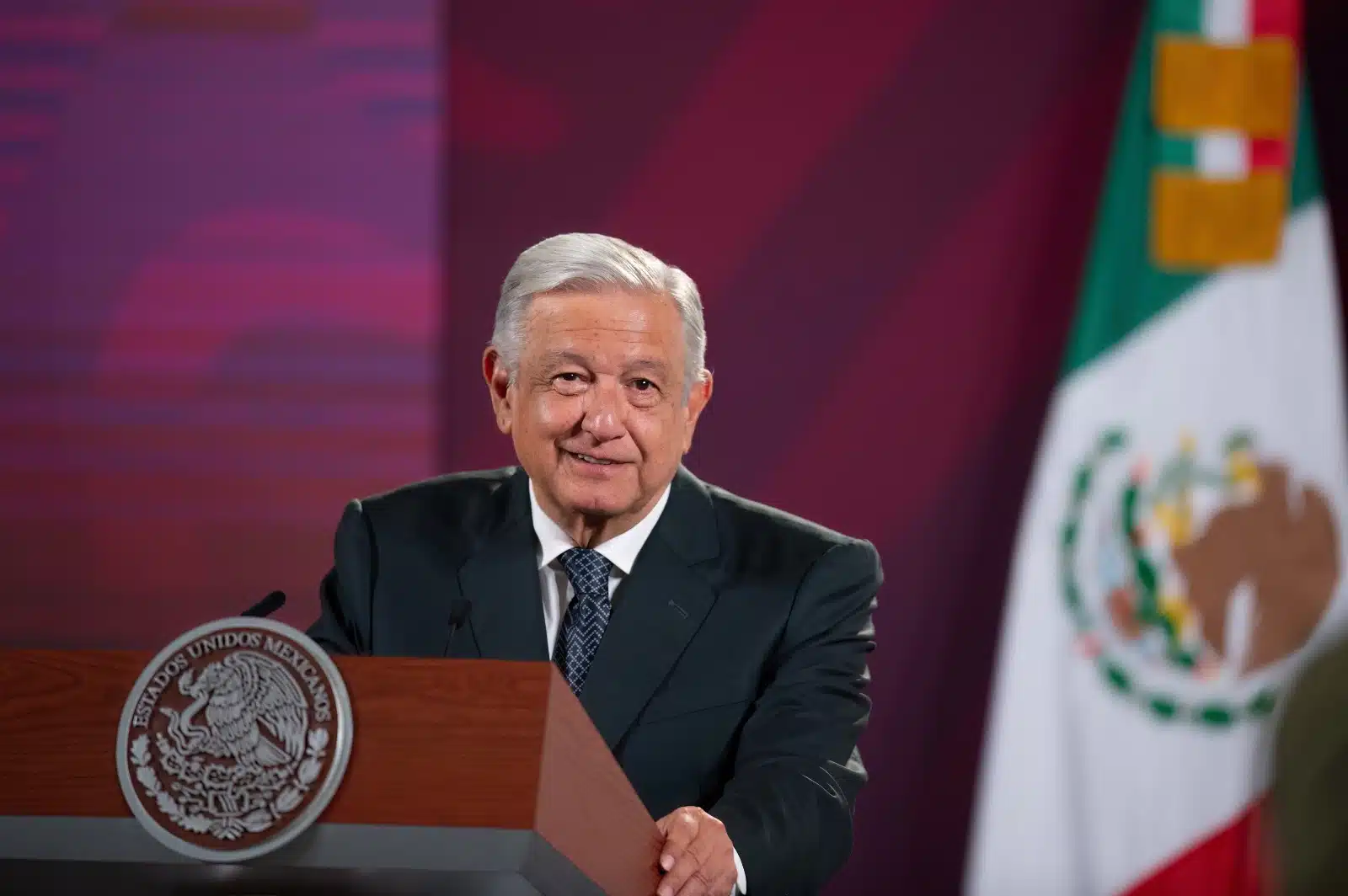 Una persona parada, un podium y la bandera de México al fondo