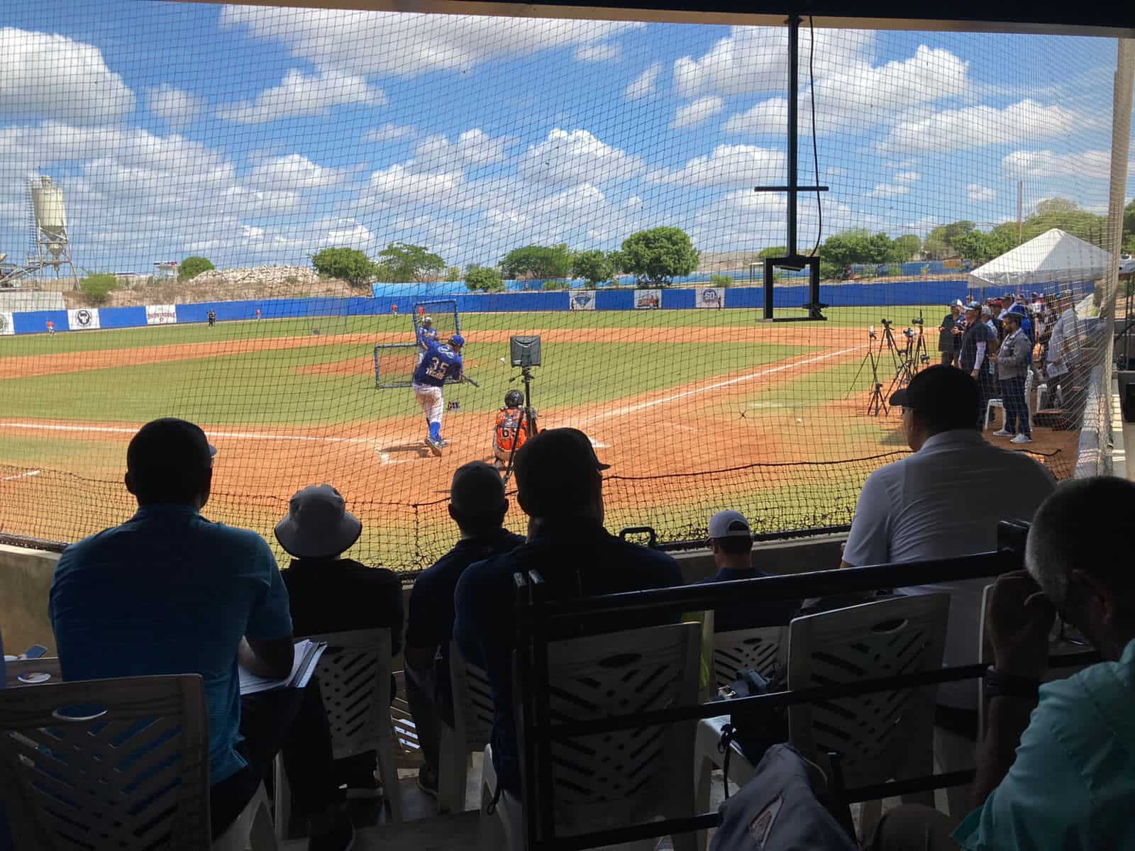 Personas sentadas y paradas, al fondo un campo de besibol con césped y tierra