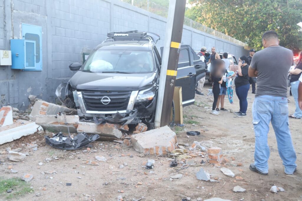 Camioneta negra chocada, poste, barda y personas al rededor