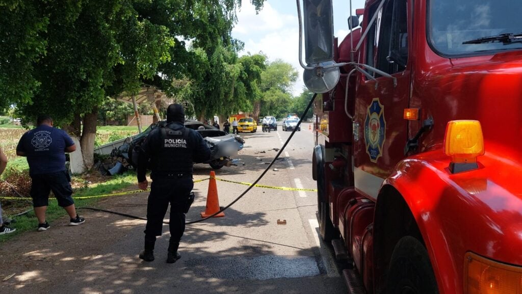 2 personas paradas, conos color anaranjados amarrados entre si con cinta amarilla delimitando un área de accidente, carro chocado, árboles, parcela, banca de fierro, una parte delantera de un camión color rojo y al fondo carros