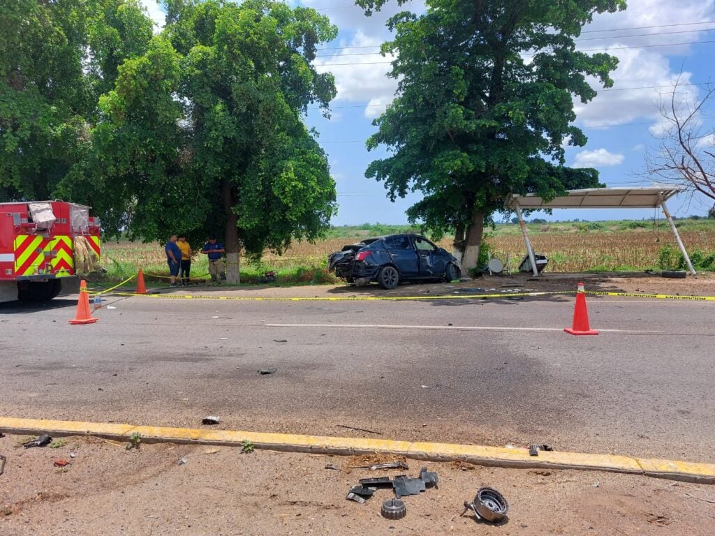 Conos anaranjados amarrados entre si con cinta amarilla delimitando un área de accidente, carro chocado, árboles, parcela, banca de fierro y la parte trasera de un camión color rojo con amarillo