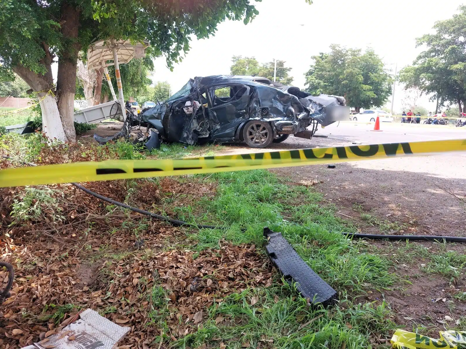 Un carro chocado, una banca de fierro, cinta amarilla delimitando el área del accidente, 1 cono color anaranjado y árboles
