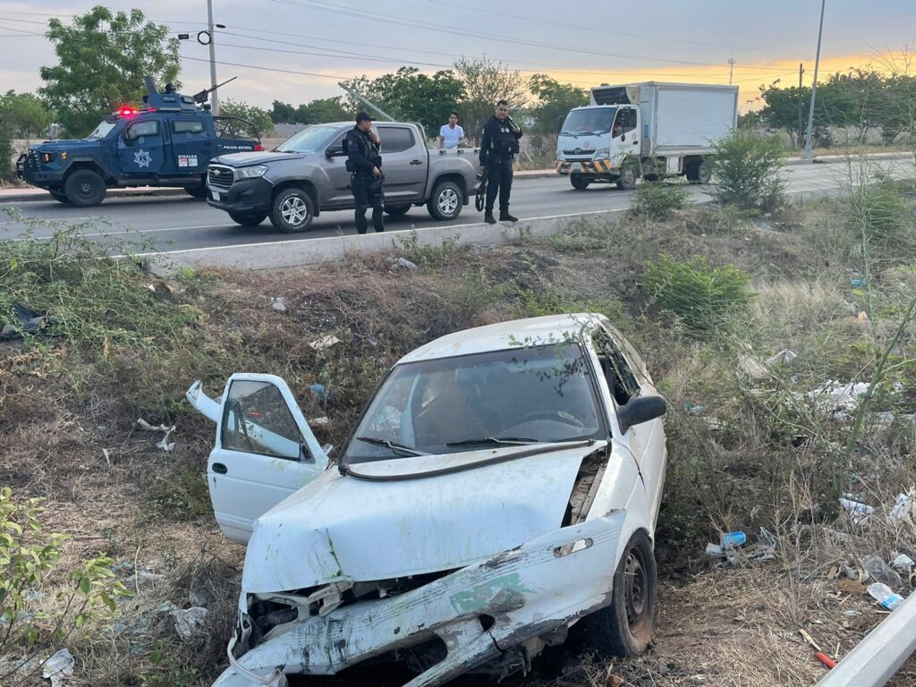 Un carro color blanco chocado del frente con la puerta del copiloto abierta, ramas, postes de luz, 3 personas mirándolo, 2 camionetas y un camión