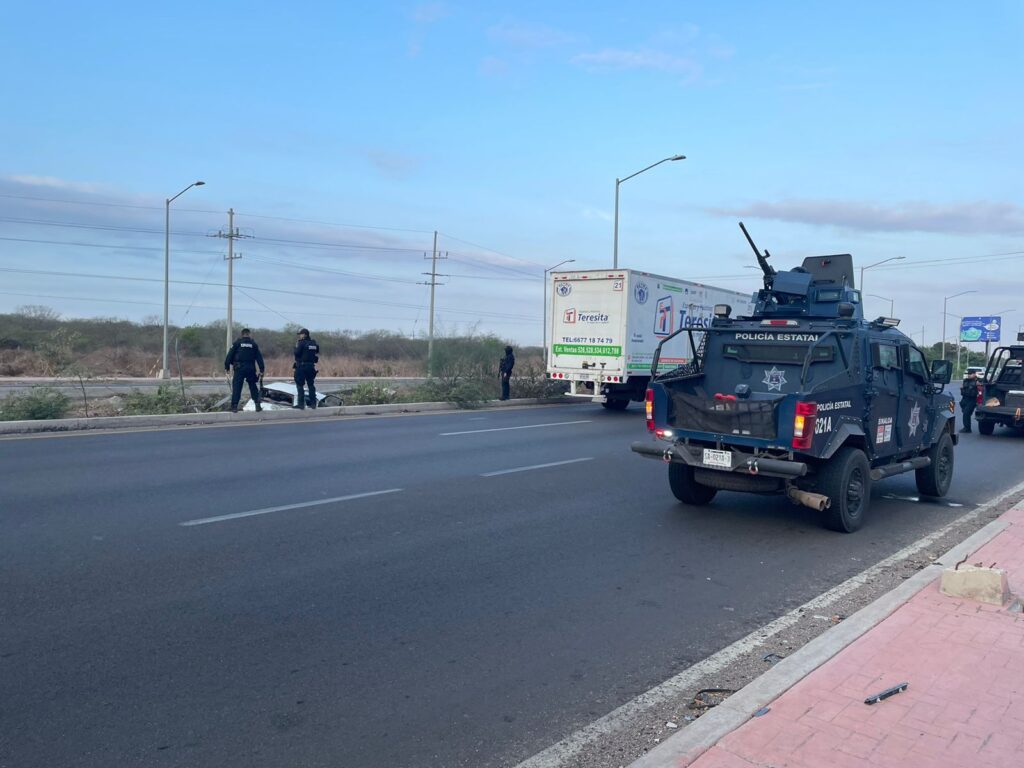 Una camioneta y parte de otra estacionadas sobre la carretera y la caja de un tráiler, postes de luz, 3 personas mirando un carro que quedó sobre el camellón