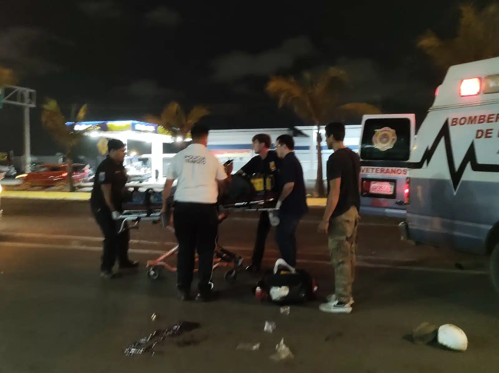 4 personas agarrando una camilla y otra parada a un lado sobre la carretera, y la parte trasera de una ambulancia con la puerta abierta, al fondo palmeras, carros y una estación de gas