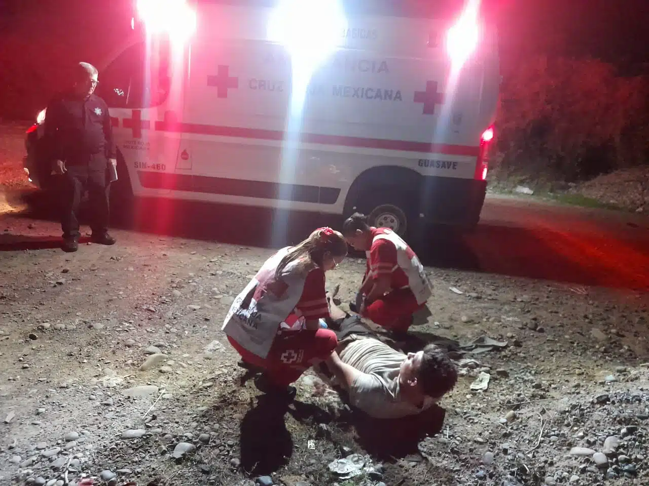 2 personas atendiendo a una en el suelo y una más mirando a un lado de una ambulancia con las luces prendidas