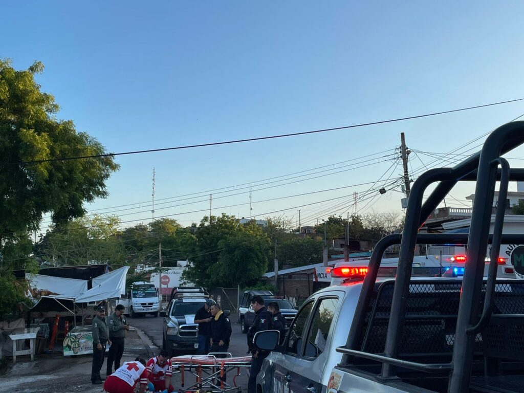 3 camionetas de la policía municipal de Culiacán, una ambulancia de la Cruz Roja y un camión sobre una calle, casas, árboles, personas y una camilla