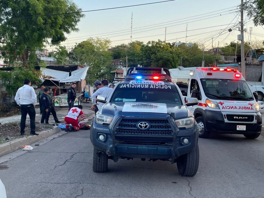 Una camioneta de la policía municipal de Culiacán, una ambulancia de la Cruz Roja y 7 personas parada mirando a una que le da auxilio a otra
