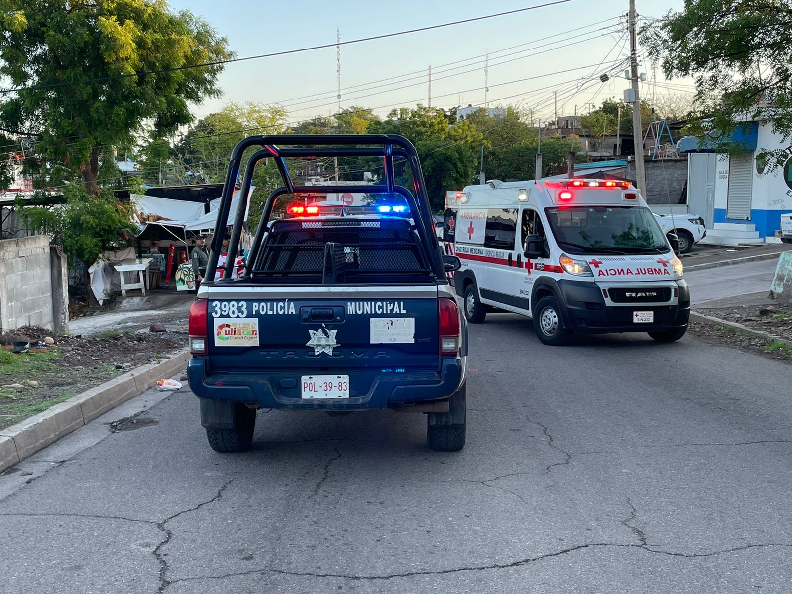Una camioneta de la policía municipal de Culiacán y una ambulancia de la Cruz Roja sobre una calle, casas y árboles