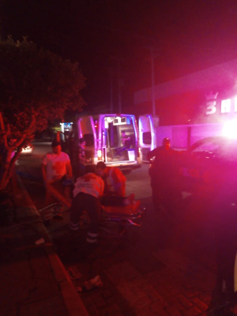2 personas ayudando a otra en una camilla, una más parada recargada sobre una camioneta mirando y una ambulancia con las puertas abiertas