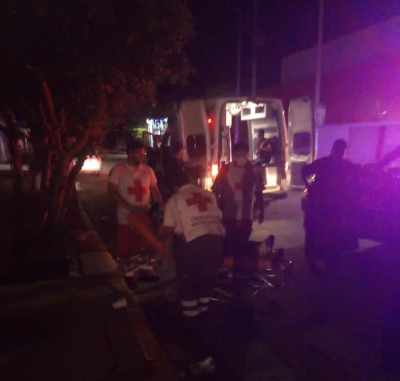 2 personas ayudando a otra en una camilla, una más parada recargada sobre una camioneta mirando y una ambulancia con las puertas abiertas