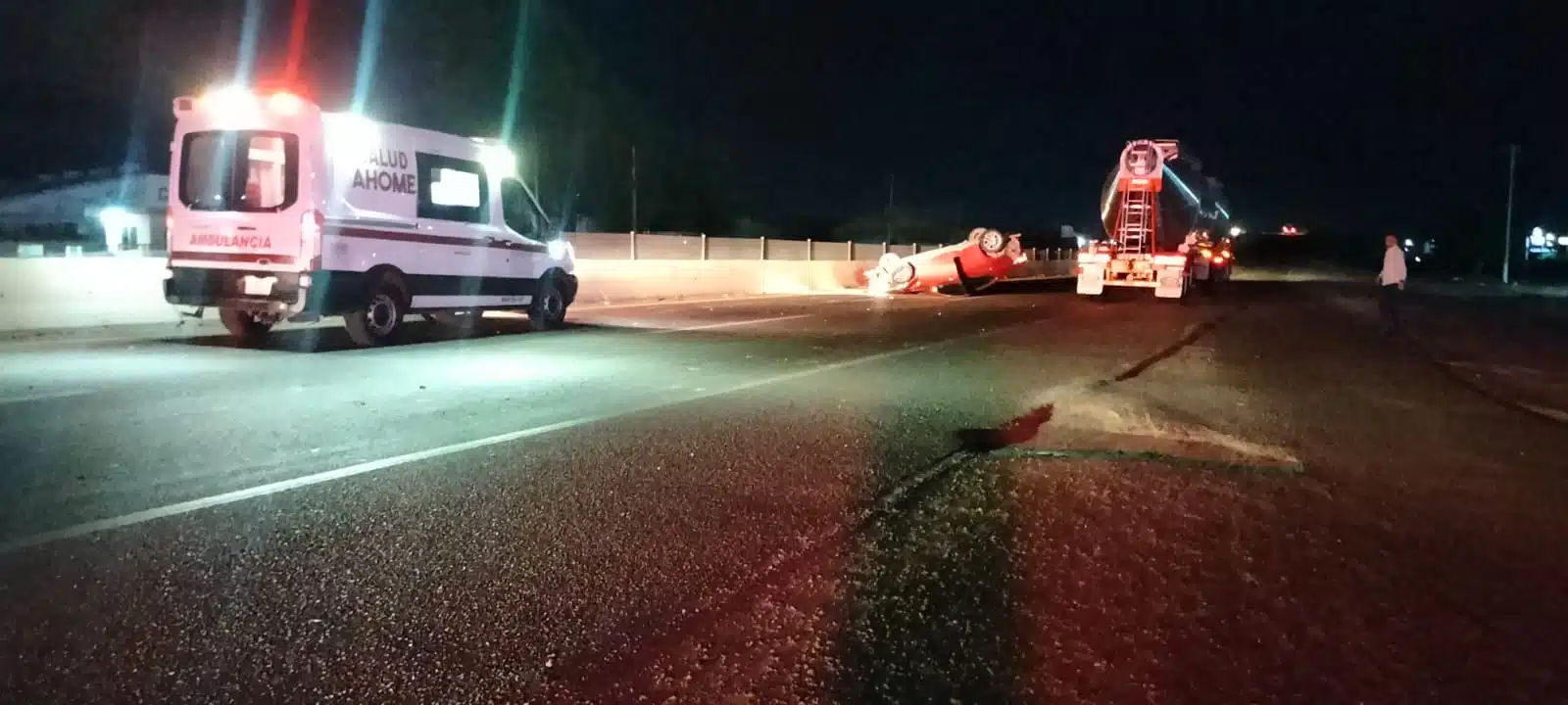 Un carro color rojo con los neumáticos hacia arriba sobre la carretera, una ambulancia con los focos prendidos y un camión