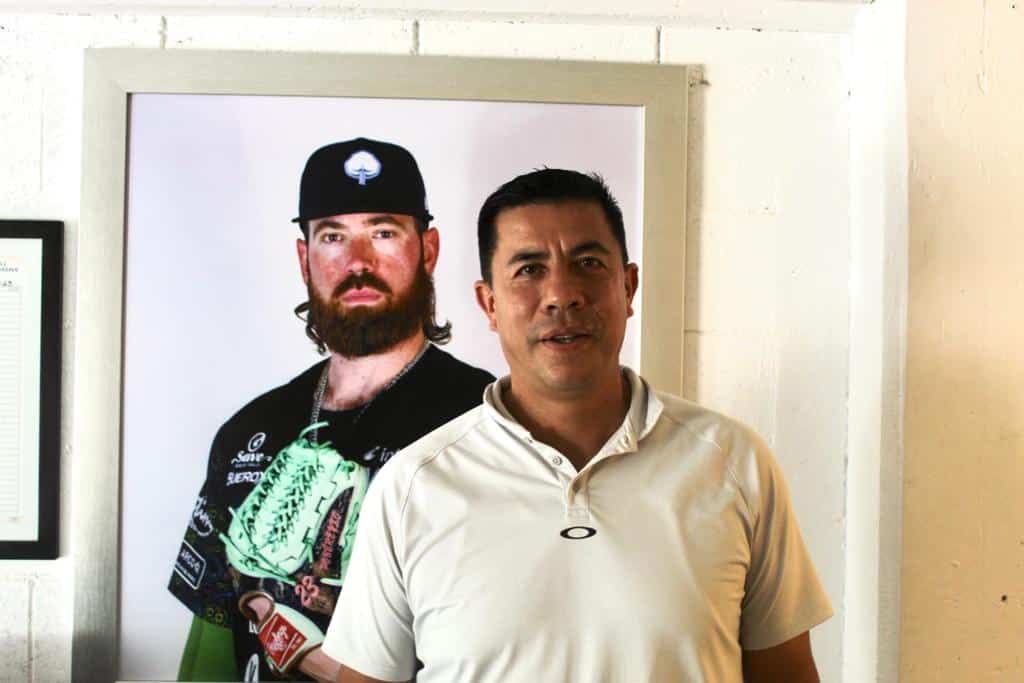 Una persona con playera blanca y atrás un cuadro con una fotografía de otra persona