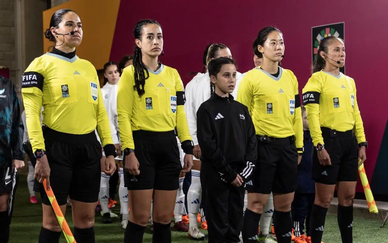 5 mujeres 4 de ellas con uniforme de árbitro color amarillo con negro y la otra de negro, al fondo más personas