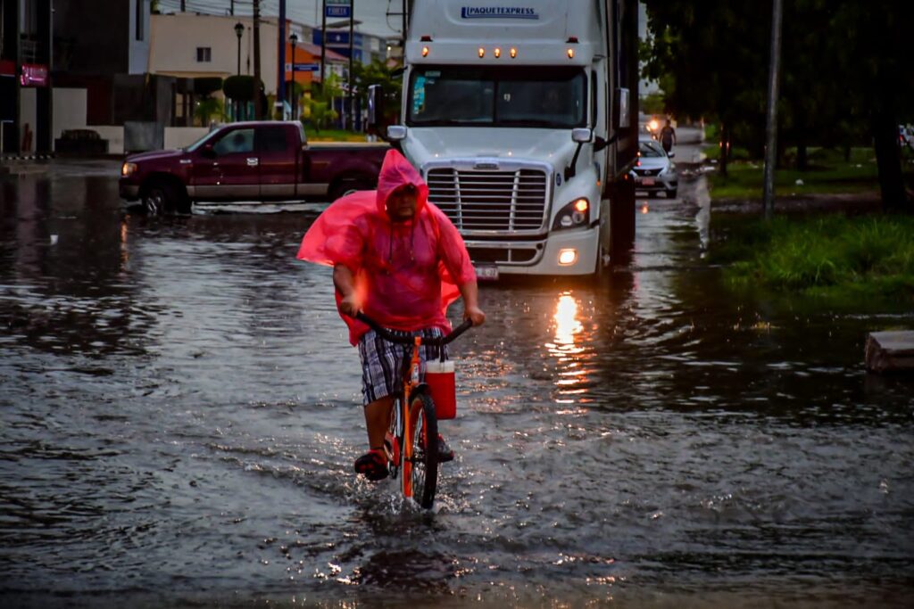 Una persona en bicicleta, calle inundada y un tráiler al fondo