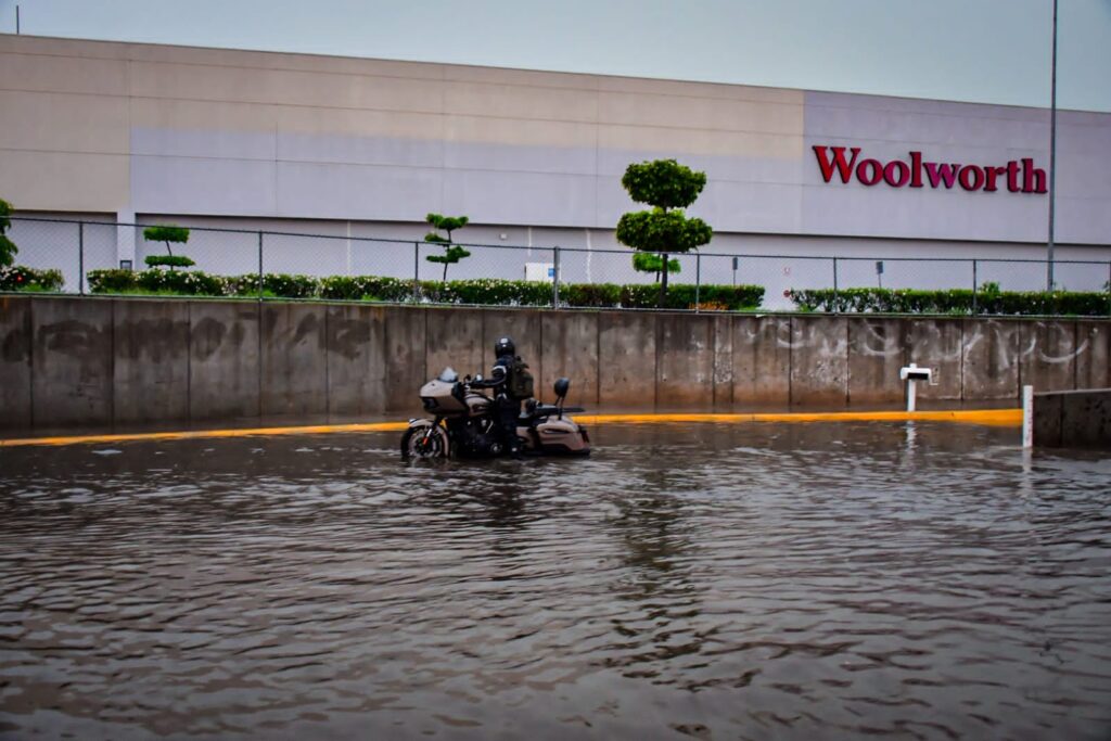 Avenida inundada, árboles, pared de un centro comercial y un hombre en moto