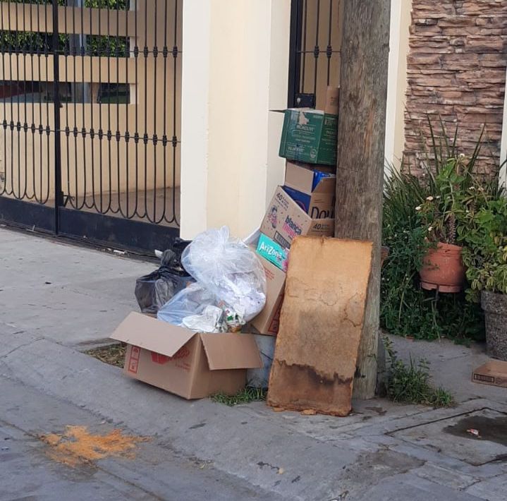 Cartones y bolsas de basura en la banqueta junto a un poste, barandal de una cochera y paredes de una casa, y plantas en maceta