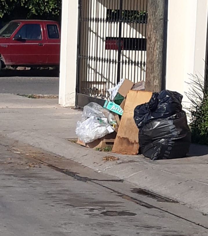 Cartones y bolsas de basura en la banqueta junto a un poste, barandal de una cochera y paredes de una casa, una camioneta color rojo al fondo, y plantas en maceta