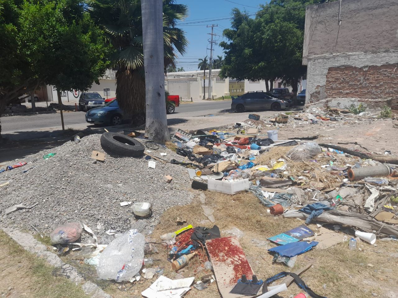 Basura, llantas y escombros tirados en un baldío, carros y árboles al fondo