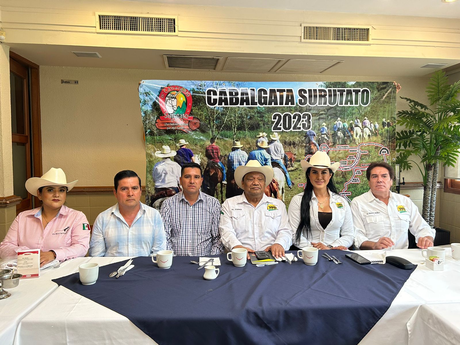 6 personas sentadas 3 de ellas con sombrero, tazas, cubiertos, una mesa con un mantel de color blanco con azul, libretas y celulares
