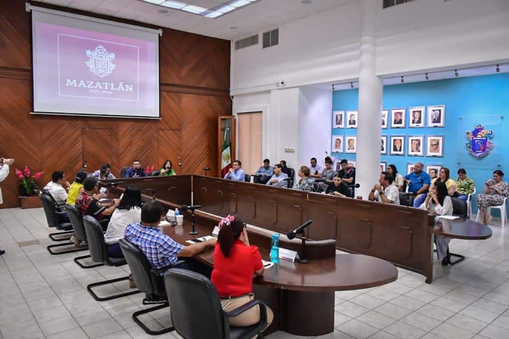 Personas sentadas, una mesa tipo escritorio ovalado sin una parte, micrófonos, cuadros, paredes de color azul, blanco y tipo madera, muros y pantalla de un proyector con letras y un logo