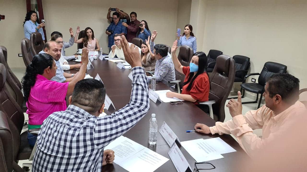 Muchas personas sentadas con la mano levantada, otras paradas, hojas sobre una mesa de color café, sillas y botellas de agua