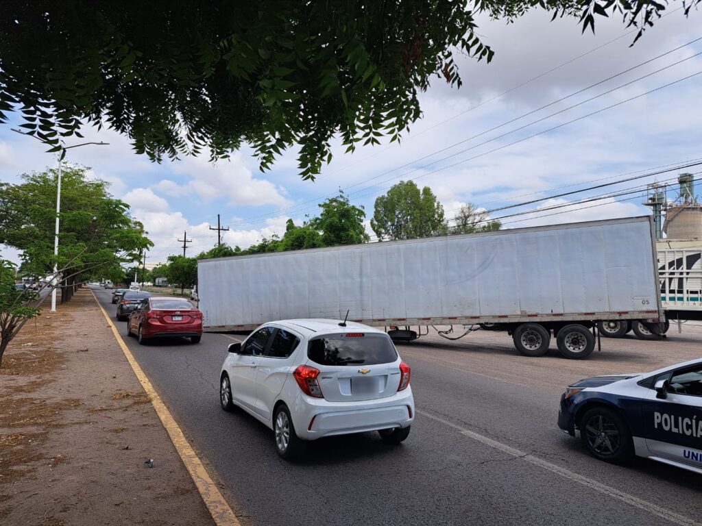 Caja de un tráiler volcada sobre una carretera, una fila de carros al lado, árboles, postes, un camellón y cielo con nubes