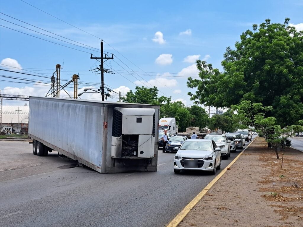 Caja de un tráiler volcada sobre una carretera, una fila de carros al lado, un camellón, árboles, postes y cielo con nubes