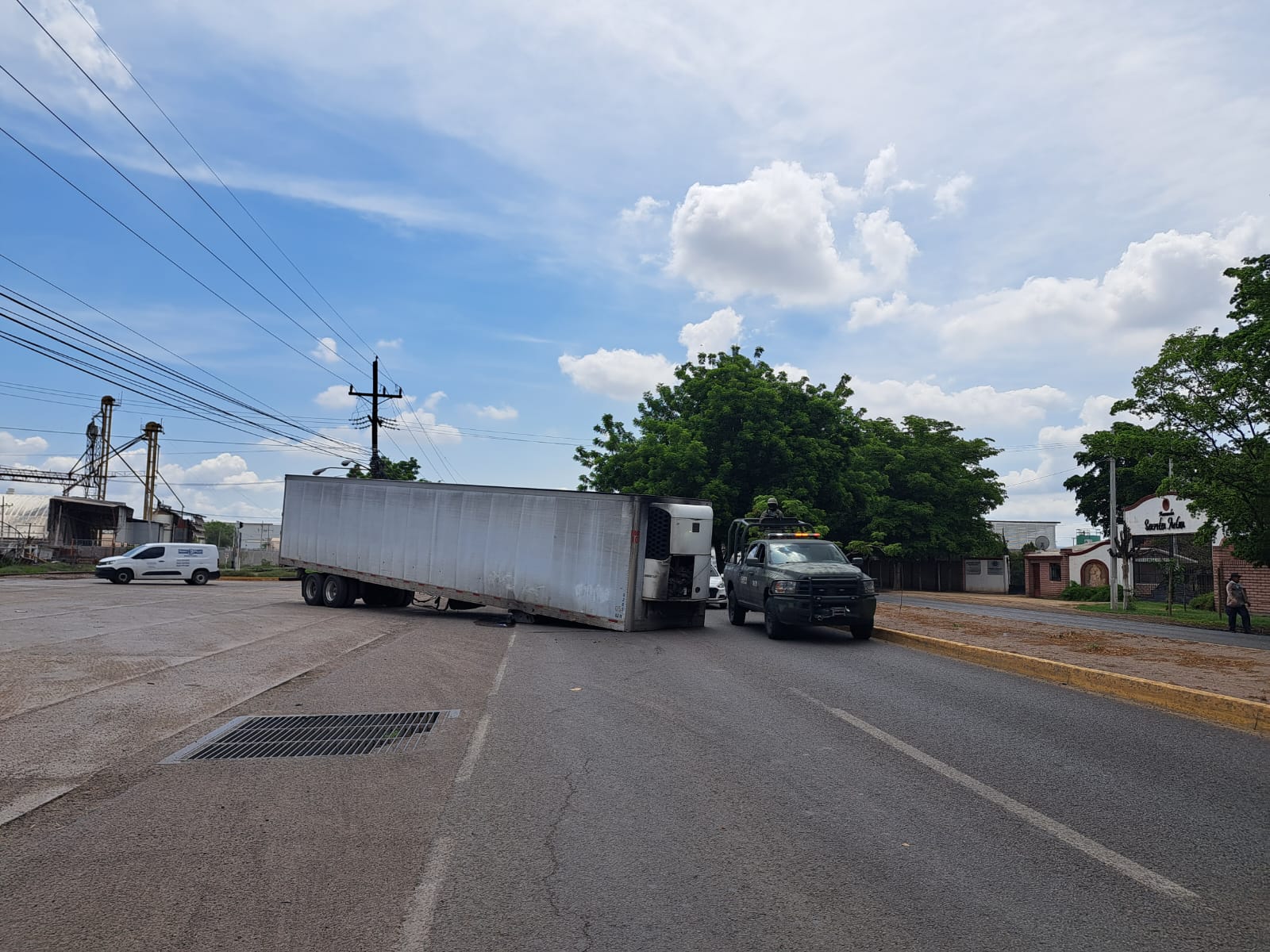 Caja de un tráiler volcada sobre una carretera, una patrulla al lado, árboles y cielo con nubes