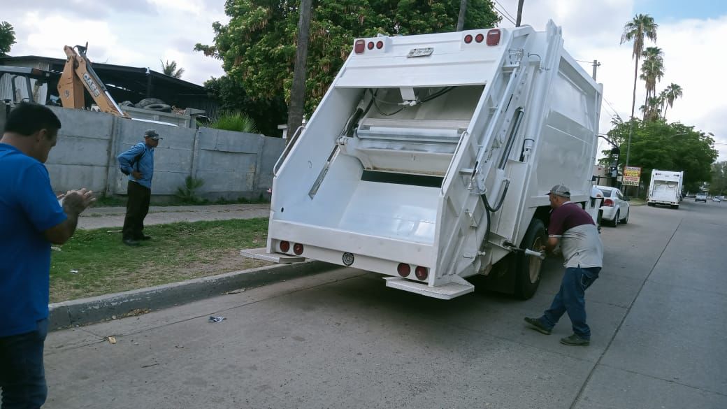 Camion de la basura blanco estacionado en una calle, personas al rededor y árboles la fondo