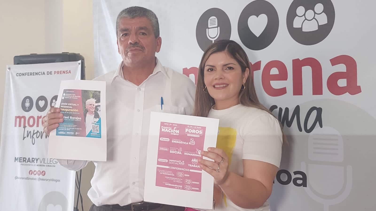2 personas posando para una foto con 2 hojas una cada uno, atrás de ellos una lona con letras de un partido político y a un lado un estante del mismo partido