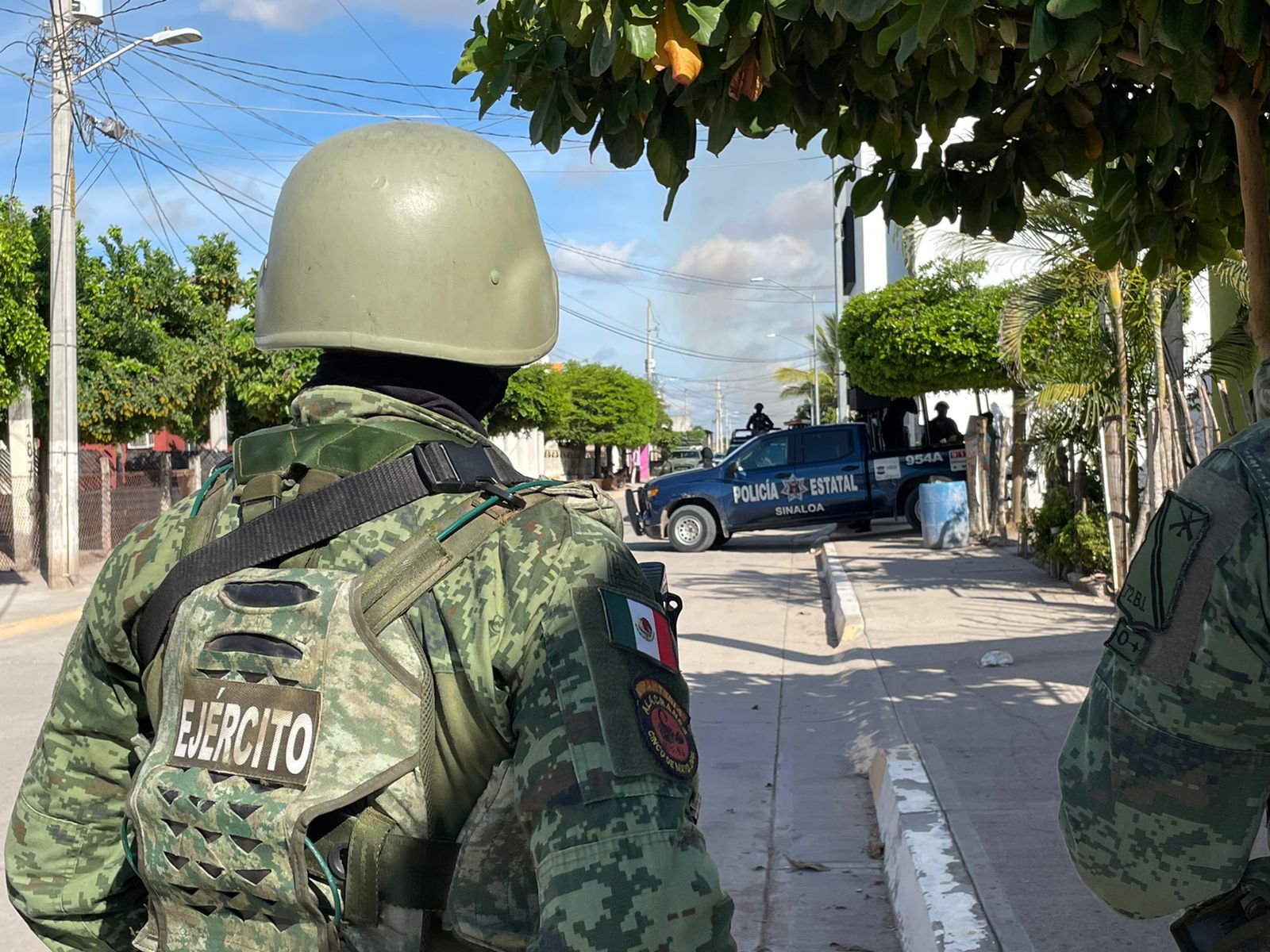 Una persona con ropa y casco color verde, una camioneta de la Policía Estatal, árboles y casas