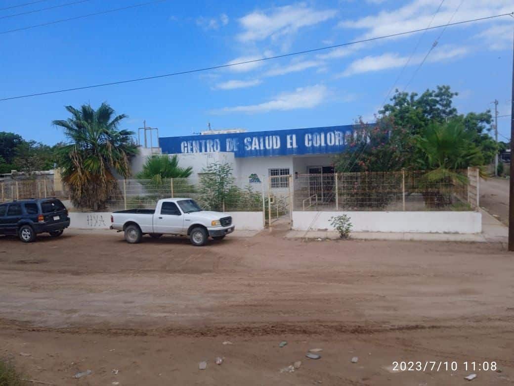 Un centro de salud, 2 camionetas estacionadas en la calle, árboles y postes