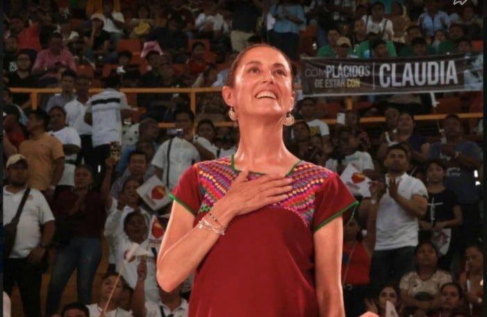 Una persona sonriendo con la mano en el corazón y más personas al fondo