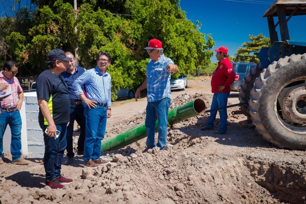 6 personas paradas en una obra, tubos, parte trasera de un tractor, la parte del frente de un carro y parte trasera de una camioneta y árboles