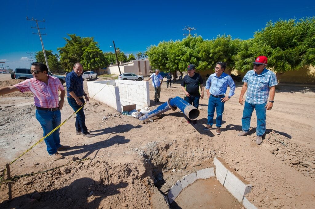 5 personas mirando una obra, otra recargada en una pared, una más al fondo, cinta amarilla árboles, camionetas y tubos