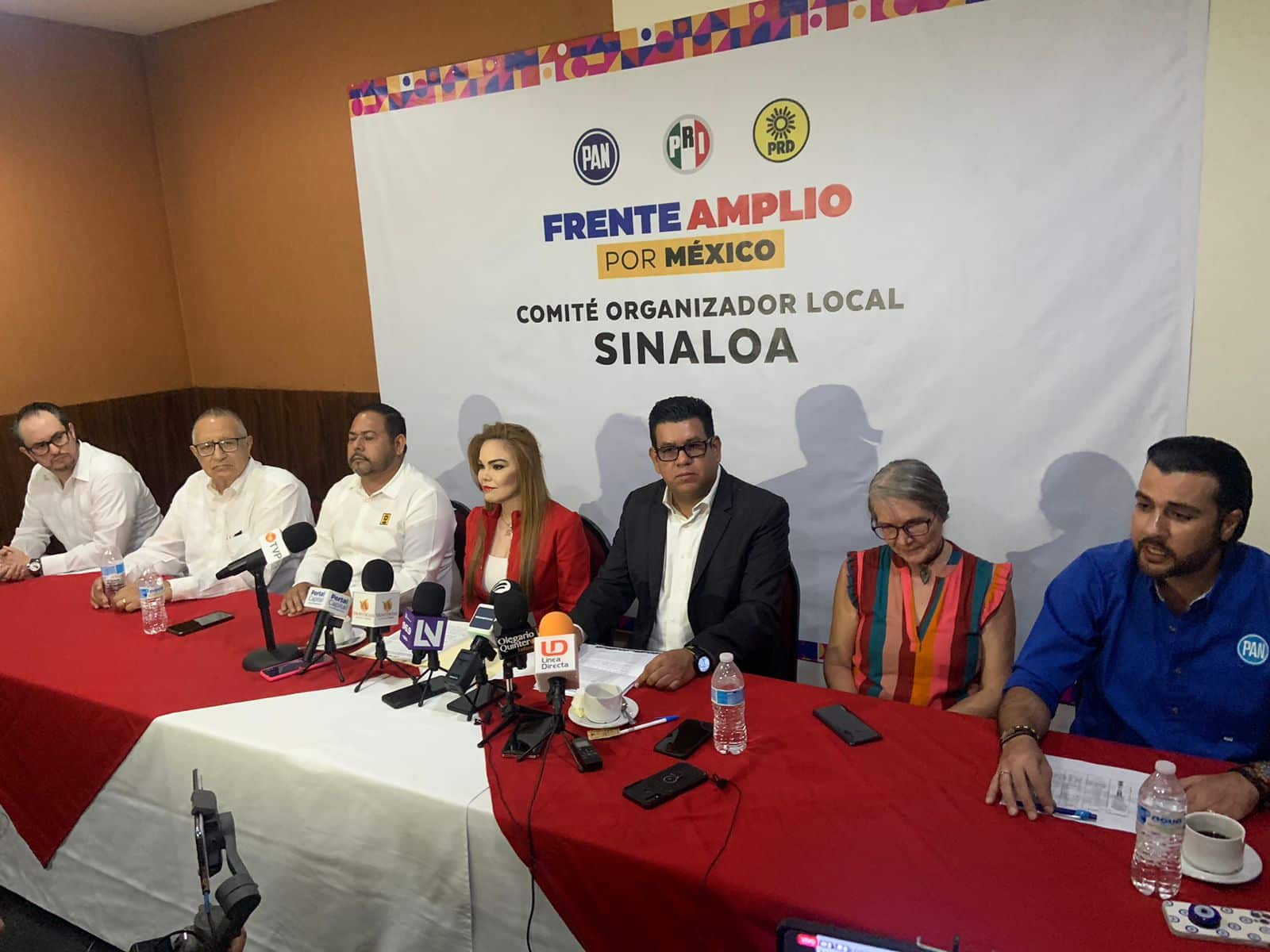 7 personas sentadas, una mesa con un mantel blanco con rojo, micrófonos, celulares, tazas y botellas de agua, al fondo una lona con letras y logotipos