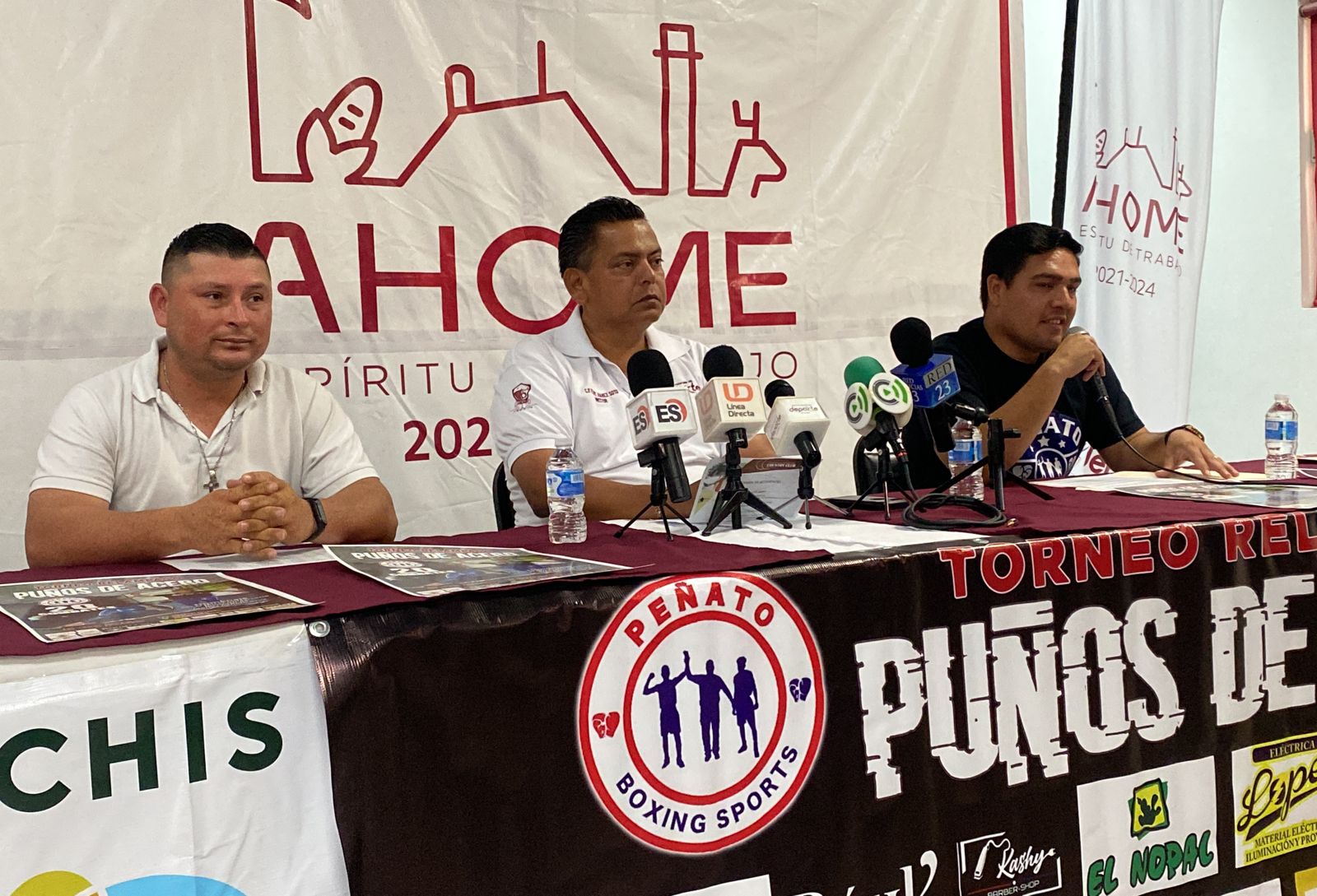 3 personas sentadas, una mesa, 5 micrófonos, aguas, carpetas, posters, sillas y una lona al fondo con letras y dibujos, otra más enfrente