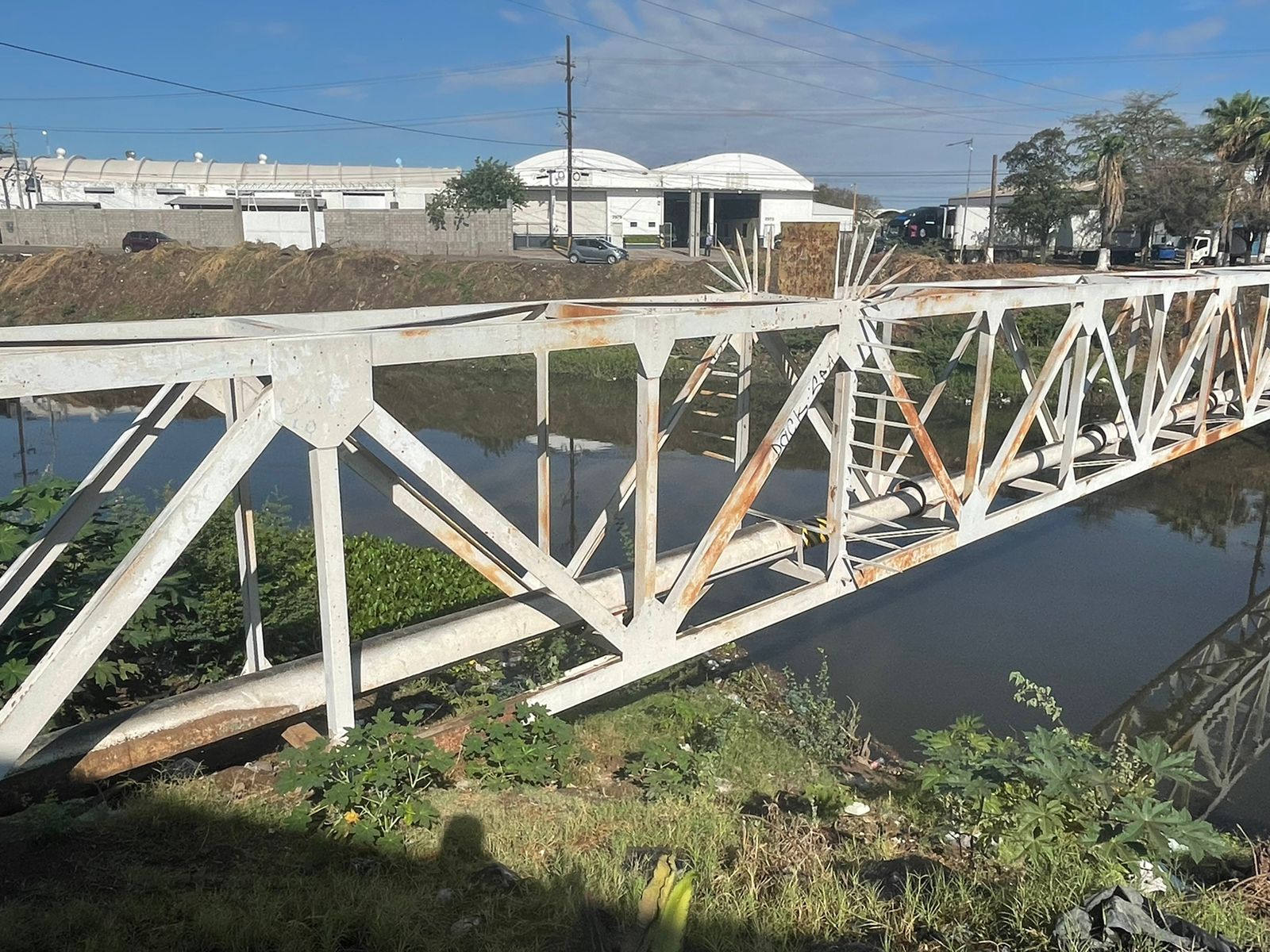 Una estructura de fierro que pasa un canal con agua, bodegas y carros al fondo