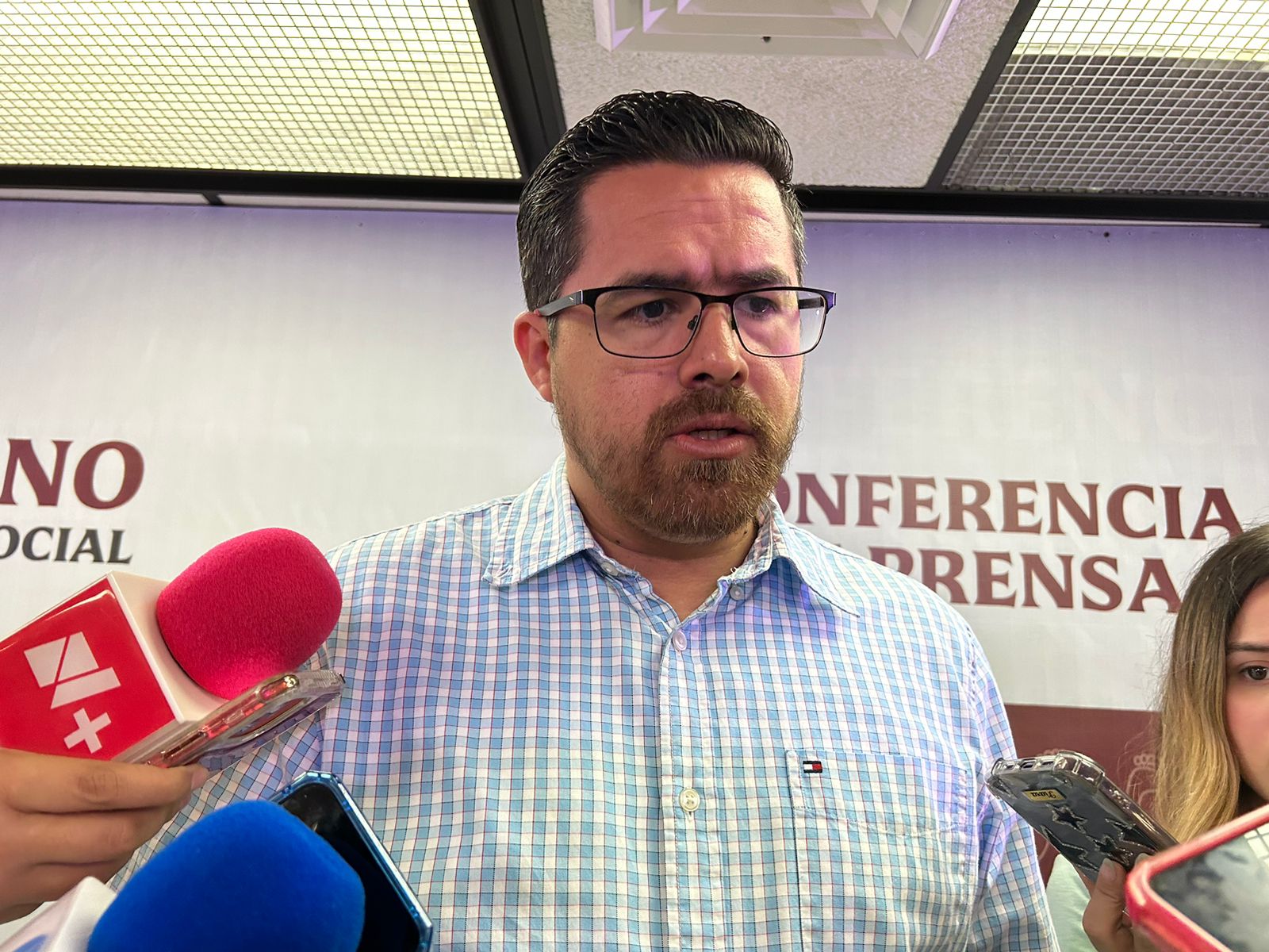 Una persona con lentes y barba hablando ante los medios de comunicación, micrófonos y celulares